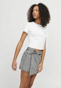 Gonna avvolgente in denim grigio con vita alta, dettaglio a pattina anteriore e due bottoni. Abbinata a una camicia aderente bianca a maniche corte.