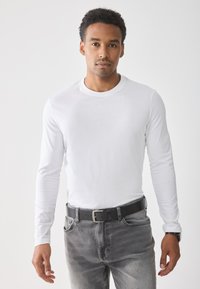 Weißes Langarmhemd aus weichem Stoff, kombiniert mit grauen Jeans. Ein schwarzer Gürtel mit einer silbernen Schnalle vervollständigt das Outfit. Schlichtes Design.