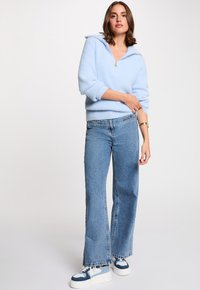 Suéter esponjoso azul claro con cuello con cremallera, combinado con unos pantalones vaqueros de tiro alto y pierna ancha en azul claro y zapatillas blancas con acentos azules.