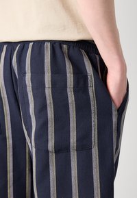 Pantalons rayés navy en tissu texturé, avec des rayures verticales blanches et gris clair, et une taille élastique. Comprend des poches latérales.