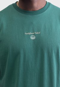 T-shirt en coton vert avec un col rond. "Redefined Rebel" brodé en rose clair au-dessus d'un design graphique simple. Texture douce.
