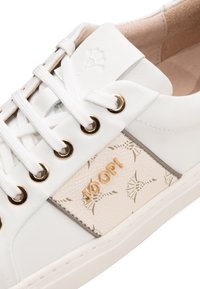 Weiße Ledersneaker mit goldfarbenen Ösen, ausgestattet mit einem strukturierten beigen Einsatz mit Muster und geprägt eingearbeitetem Logo. Klassisches Schnürdesign.