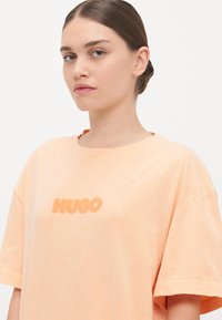 Růžová oversized tričko vyrobené z jemné látky, s texturovaným logem "HUGO" uprostřed a širokým kulatým výstřihem.