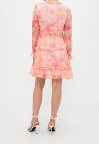 Roze, doorschijnende jurk met bloemenpatroon, lange mouwen, geplooide lagen en een getailleerde taille. Gecombineerd met witte sandalen met hoge hakken.