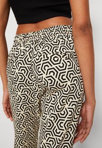 Crème-kleurige broek met een geometrisch zwart en crème patroon, voorzien van een elastische tailleband en zijzakken. De stof ziet er glad uit.