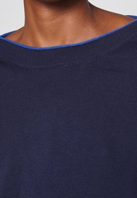 Gros plan sur le col d'un pull en maille bleu foncé avec une fine bordure bleu roi autour du cou et du haut de la poitrine d'une personne.