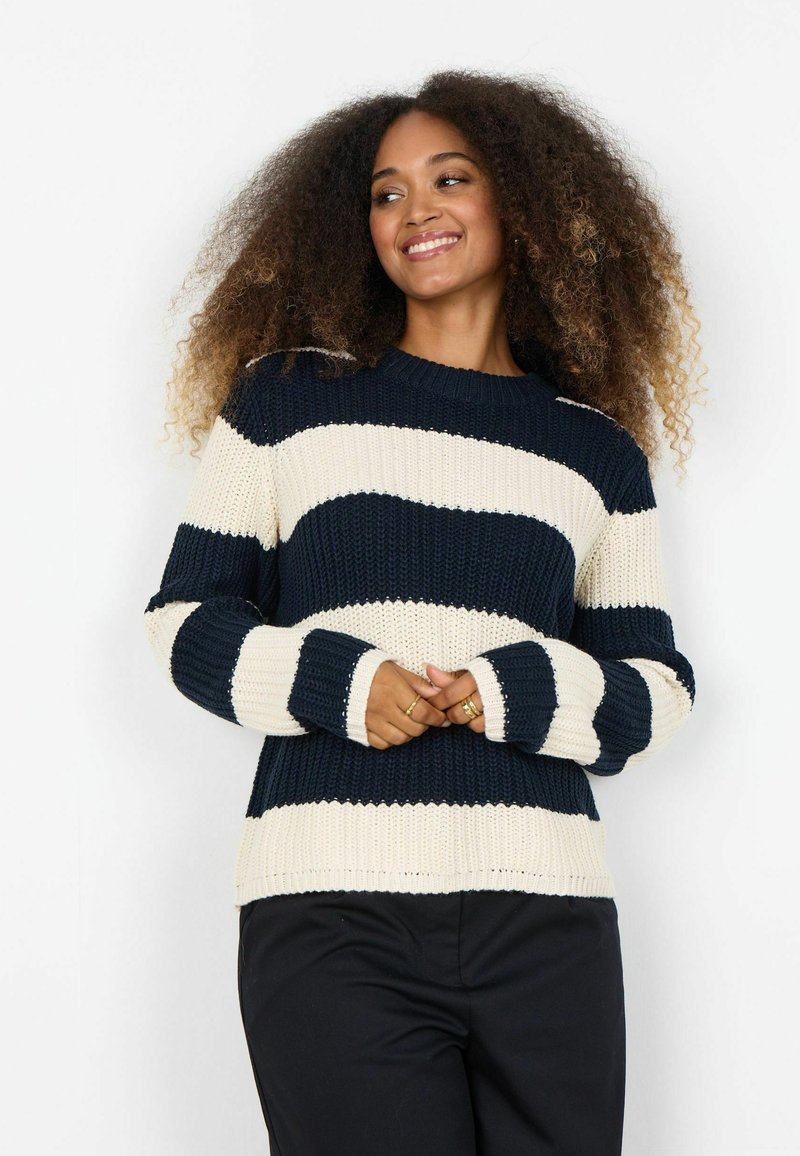 Frau mit lockigem Haar, die lächelt, trägt einen dicken Pullover mit horizontalen marineblauen und weißen Streifen sowie schwarze Hosen, die Hände vor dem Körper verschränkt.