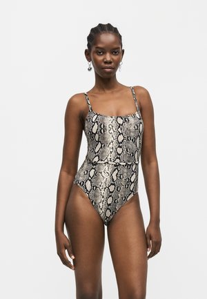 Naine, kes kannab varrukateta ülikonda, millel on madulnaha muster ja õhukesed rihmad, seisab ühevärvilise valge tausta ees.