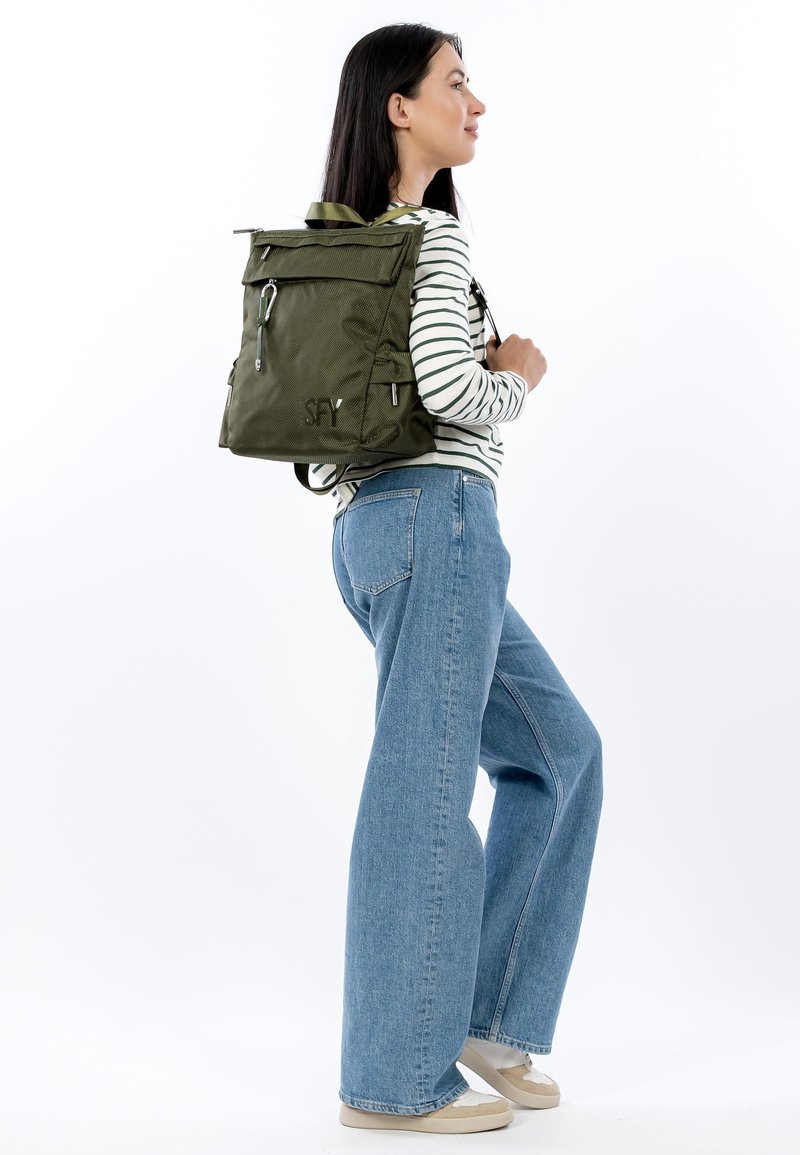 Oliviroheline tekstuuriga seljakott, millel on pealmine klapp, eesmine tõmblukuga tasku ning reguleeritavad rihmad. Sobitatud triibulise topi ja laiade teksadega.