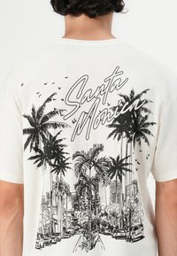 T-maica krem-v boji s črnim grafičnim dizajnom palm, mestne silhuete in besedilom "Santa Monica" v pisani pisavi.
