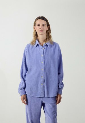 Frau trägt ein locker sitzendes, geripptes lavendelfarbenes Hemd mit Knöpfen und passende Hose, steht vor einem schlichten weißen Hintergrund.