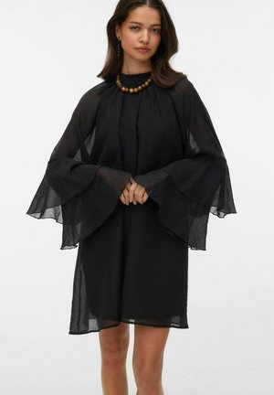 Femme portant une robe noire transparente avec de larges manches cloche et un collier de perles en bois, debout devant un fond blanc uni.