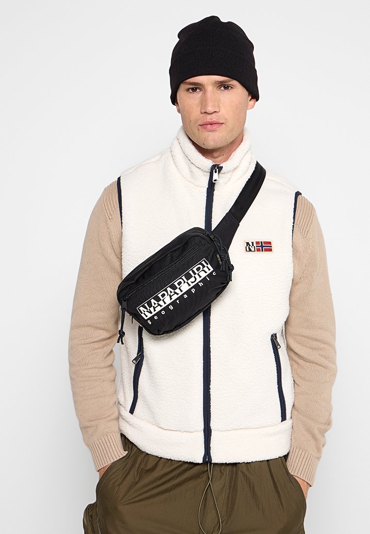 Mężczyzna w czarnej czapce beanie, białej polarowej kamizelce na beżowym swetrze, oliwkowych spodniach i czarnej torbie na ramię z białym logo "Napapijri".