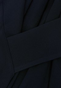 Cinturón de tela azul marino oscuro atado alrededor de una prenda texturizada a juego en azul marino oscuro con pliegues superpuestos.