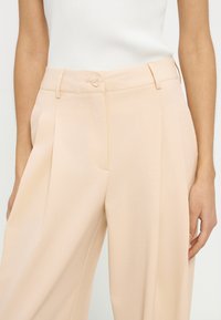 Pantalon beige taille haute à jambes larges avec plis devant et fermeture par boutons, porté avec un haut sans manches côtelé blanc.