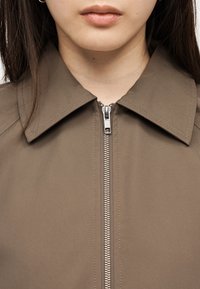 Κοντινό πλάνο ενός ατόμου που φοράει καφέ ένδυμα με γιακά και ασημένιο φερμουάρ κλεισμένο μέχρι πάνω, ορατά τα χείλη και το σαγόνι, με μακριά σκούρα μαλλιά.