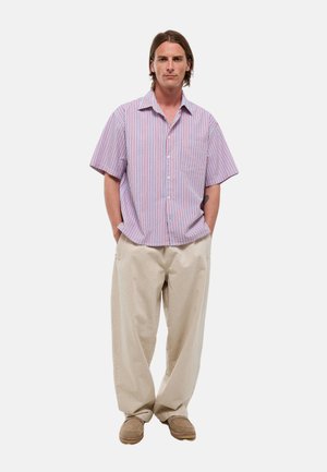 Homme debout les mains dans les poches, portant une chemise à manches courtes rayée rouge et bleue, un pantalon beige à jambes larges et des chaussures marron.