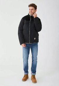 Čierna puffková bunda s vysokým golierom, viditeľným šitím a prednými gombíkmi. Nosená s vyhrnutými modrými džínsami a hnedými čižmami.