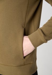 Hand delvis inskjuten i sidofickan på en olivgrön långärmad sweatshirt med synliga sömdetaljer vid ärm- och nederkant.