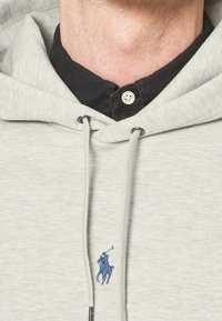 Szara bluza z kapturem i kieszenią typu kangur. Na froncie niebieskie haftowane logo z jeźdźcem konnym. Pod spodem znajduje się czarna koszula z kołnierzykiem.