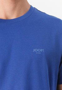 T-shirt in cotone blu con scollo rotondo e maniche corte. Presenta un piccolo logo "JOOP! JEANS" stampato in blu chiaro sul davanti.