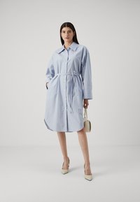 Abito da camicia a righe blu con colletto, maniche lunghe e cintura con lacci. Presenta una chiusura con bottoni e tasche. Indossato con tacchi a punta beige.