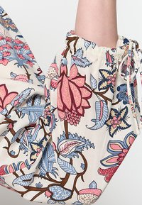 Blumenmusterstoff in Beige, mit großen rosa und blauen Blumen sowie grünen Blättern. Gummierte Ärmelbündchen und Kordelzugdetails.