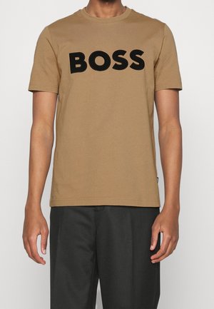 Homme portant un t-shirt beige à manches courtes avec l'inscription "BOSS" en grandes lettres noires, associé à un pantalon gris foncé, sur un fond uni.