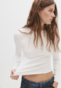 Fehér, hosszú ujjú felső, amely illeszkedő kialakítású, könnyű anyagból készült. Kerek nyakkivágással és enyhén cropped alsó résszel rendelkezik.