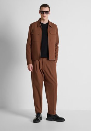 Hombre vestido con chaqueta y pantalones marrón oscuro, suéter negro, zapatos de vestir negros y grandes gafas de sol oscuras, de pie contra un fondo claro y liso.