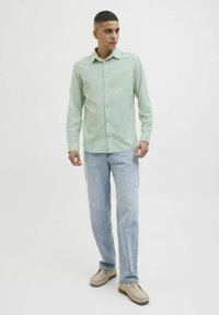 Cămașă lungă cu mâneca lungă, de culoare verde deschis, cu închidere cu nasturi la partea din față, asociată cu blugi de culoare albastru deschis și pantofi casual bej. Design simplu al gulerului.