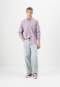 Junger Mann, der mit einer Hand in der Tasche steht, trägt ein helllila Button-up-Hemd, hellblaue Jeans und rote Sneakers vor einem weißen Hintergrund.