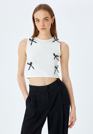 Top corto blanco de canalé con acentos de lazo negro en los hombros. Combinado con pantalones negros de talle alto, que destacan un ajuste elegante y un diseño limpio.