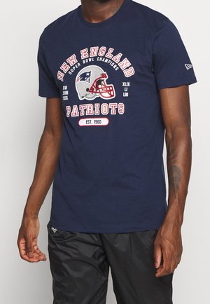 Mężczyzna w granatowej koszulce New England Patriots z grafiką kasku, tekstem mistrza Super Bowl i czarnych spodniach na jednolitym tle.