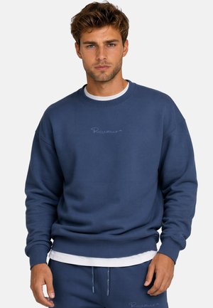 Giovane uomo con capelli corti castani, che indossa una felpa blu navy e pantaloni della tuta abbinati, su uno sfondo chiaro e uniforme.