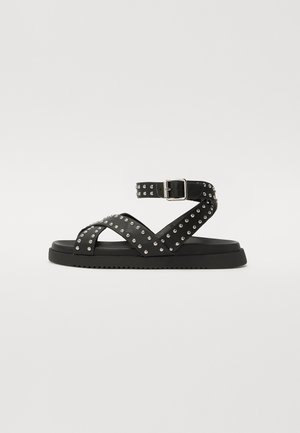 Sandal nero con cinturini borchiati, fibbia regolabile, suola piatta e fondo texturizzato per una buona aderenza. Design elegante con elementi superiori incrociati.