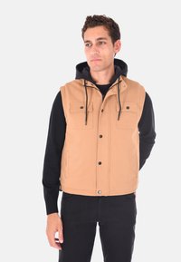 Gilet trapuntato beige con cappuccio nero, caratterizzato da bottoni a pressione e tasche frontali. Indossato sopra una maglietta nera a maniche lunghe, con una finitura testurizzata.