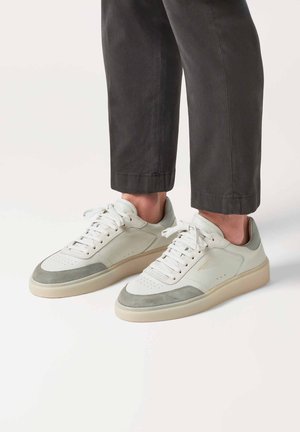Białe i szare niskie sneakersy z gładką skórzaną i zamszową cholewką, perforowanym noskiem, grubą beżową podeszwą i płaskimi białymi sznurówkami.