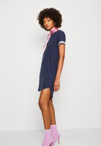 Robe T-shirt marine à manches roulées, fentes sur les côtés, accompagnée d'une écharpe à motifs rose et de chaussettes à talons texturées roses.