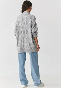 Szary sweter dziewiarski z teksturowanym wzorem w warkocze, w luźnym kroju, z podwiniętymi mankietami, zestawiony z jasnoniebieskimi spodniami o szerokich nogawkach i białymi butami.