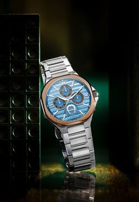 Sudraba nerūsošā tērauda pulkstenis ar zilu, teksturētu ciparnīcu un trim apakšciparnīcām. Iezīmes ietver brūnu rāmi, izteiksmīgas stundu marķējumu un saites aproci.