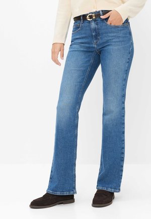 Mujer con jeans azules estilo bootcut, cinturón negro, suéter beige y mocasines marrón oscuro, de pie con una mano en el bolsillo.