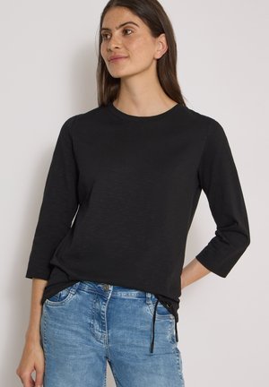 Femme aux longs cheveux foncés portant un haut noir à manches trois-quarts et un jean bleu, debout devant un fond clair uni.