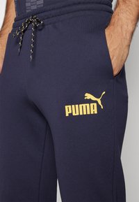 Joggers marinos de tejido suave que presentan un logo amarillo de Puma y un cordón con detalles en negro y amarillo. Equipados con bolsillos laterales.