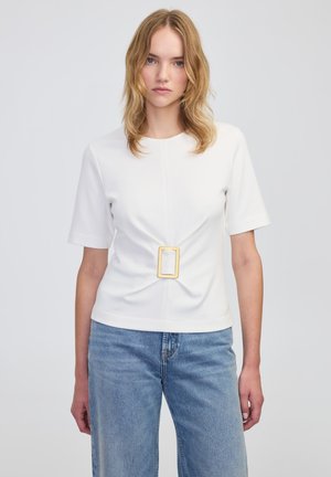 Witte korte mouw top met een centrale knoopdetail en een rechthoekige gele gespaccent. Gemaakt van een gladde stof, gecombineerd met lichtblauwe jeans.