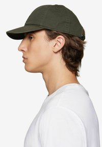 Olive green cotton baseball cap with a curved brim, adjustable back strap, and subtle branding. Worn by a person in a white t-shirt.