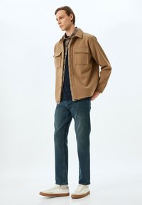 Ruskea fleece-takki, jossa on kaksi rintataskua, checkered-paitojen päällä, yhdistetty tummiin farkkuihin ja valkoisiin tennareihin, joissa on ruskeat pohjat.