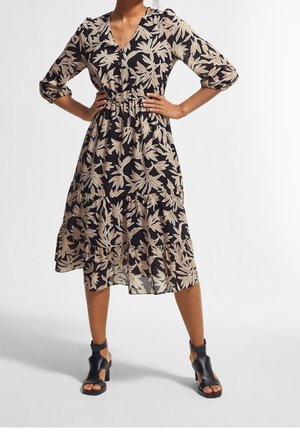 Femme portant une robe midi noire avec des motifs de feuilles beiges, manches trois-quarts, col en V, et des sandales à talons ouvertes noires, debout les mains sur les hanches.