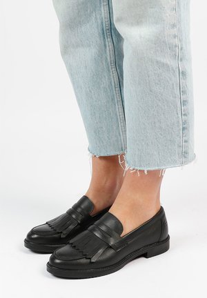 Zwarte leren loafers met franjes, platte zolen en ronde neuzen, gedragen met opgerolde lichtblauwe denim jeans.