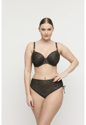 Vrouw met teruggekamd haar draagt een donkere beha met luipaardprint en een high-waisted bikini, poseert met één hand op haar heup.